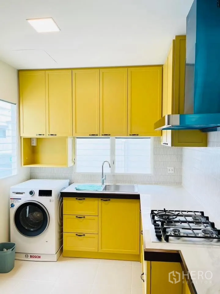 บ้านเดี่ยว for rent in Bang Phli, Samut Prakan - ครัวรูปตัวแอลตู้สีเหลือง พร้อมเครื่องซักผ้า อ่างล้างจาน และเตาแก๊ส.