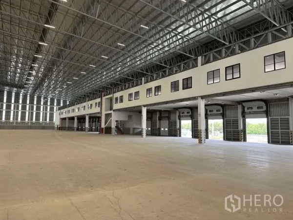 Factory for rent or sale in Samut Prakan - Vast warehouse interior with high steel truss roof, mezzanine offices and multiple roller shutters.