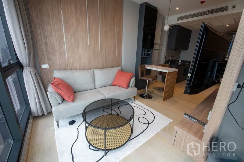 Condominium for rent in Din Daeng, Bangkok - Cozy lounge with grey sofa, orange cushions, round coffee table, wood accent wall and large windows.