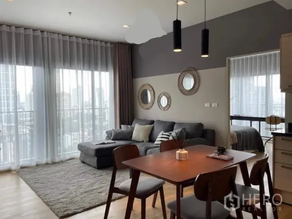 Condominium for rent in Watthana, Bangkok - Open-plan living and dining area with floor-to-ceiling windows, L-shaped sofa, and a city-view balcony.