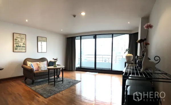 Condominium for rent in Khlong Toei, Bangkok - Spacious lounge opening to a curved glass balcony with city skyline view and a wicker sofa.
