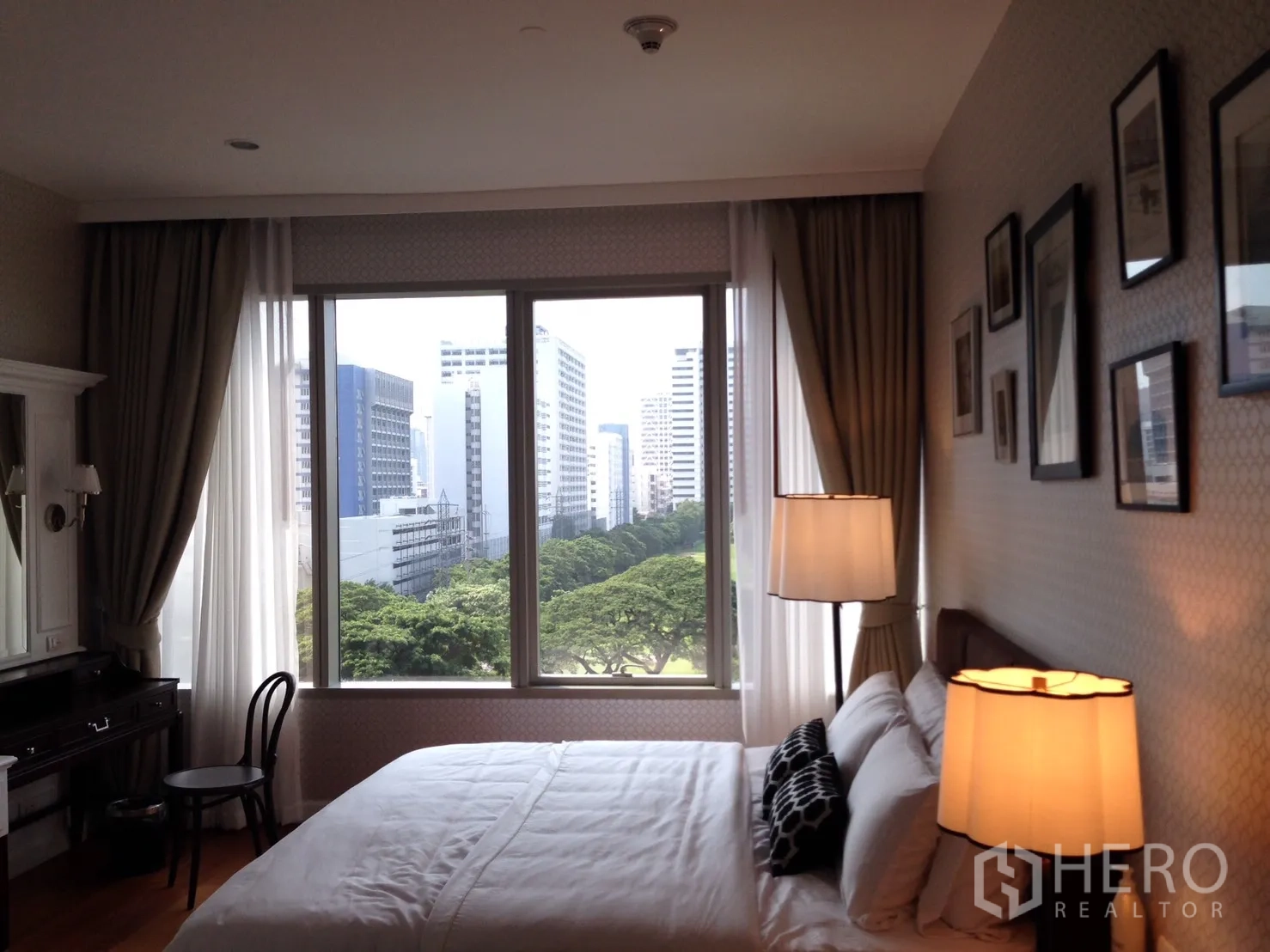 公寓 出租或出售在Pathum Wan, Bangkok - 卧室配落地窗与床头灯，可欣赏葱郁公园与城市天际线。