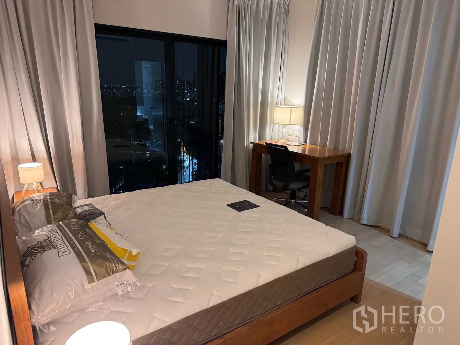 公寓 for rent or sale in Watthana, Bangkok - 主卧配大床与床头灯，窗边工作桌与座椅，推拉玻璃可见城市夜景。