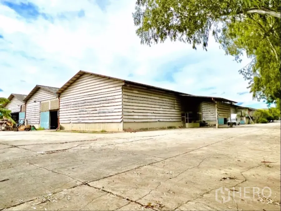 Factory for rent in Sai Noi, Nonthaburi - Wide exterior view of multi-bay factory with corrugated siding and a large concrete yard shaded by trees.