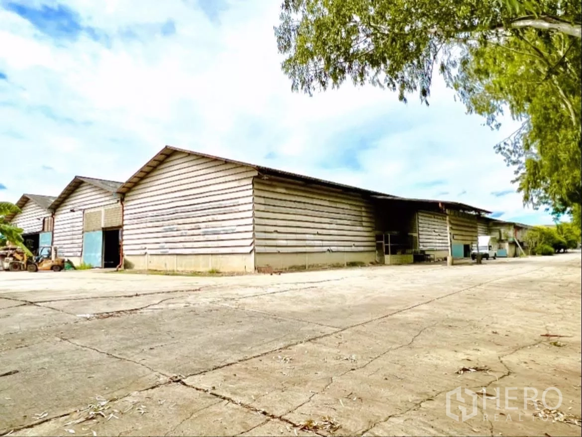 Factory for rent in Sai Noi, Nonthaburi - Wide exterior view of multi-bay factory with corrugated siding and a large concrete yard shaded by trees.