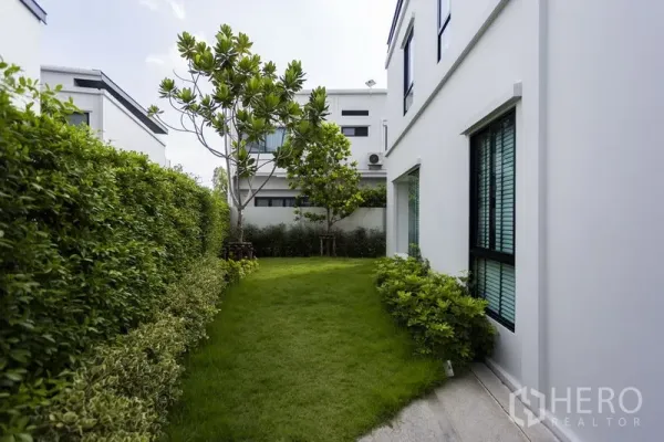 Townhouse for rent in Samut Prakan - Private side garden with lush lawn, trees and hedge beside the white townhouse.