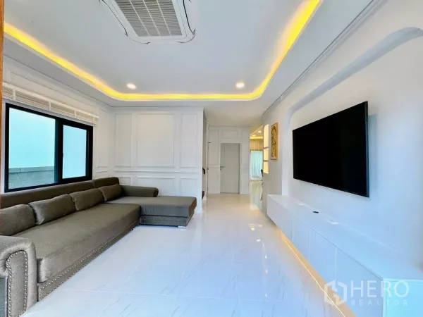 独立式住宅 出租在Bang Phli, Samut Prakan - 采光充足的客厅，L形沙发、白色地砖与灯槽天花和大电视墙。