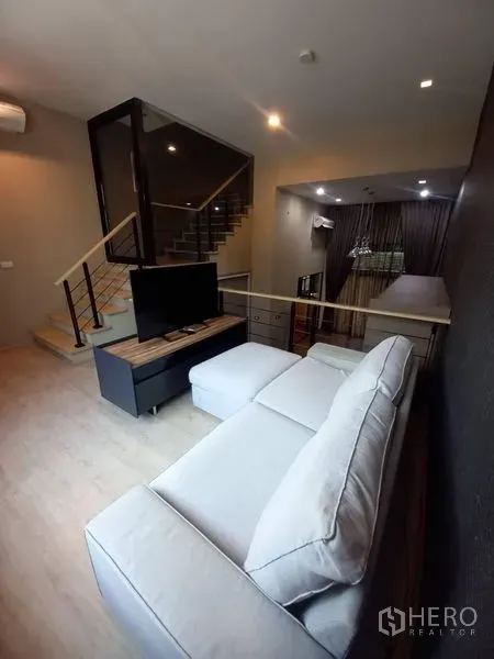 Townhouse for rent in Suan Luang, Bangkok - Open-plan living room with L-shaped sofa, TV console, wood floors and a staircase with glass balustrade.