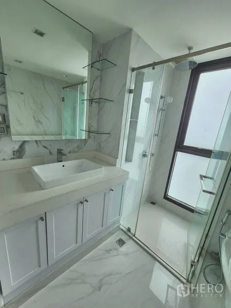 Condominium for rent in Bang Rak, Bangkok - Vanity with large mirror and basin beside a clear glass shower in a marble‑style bathroom.