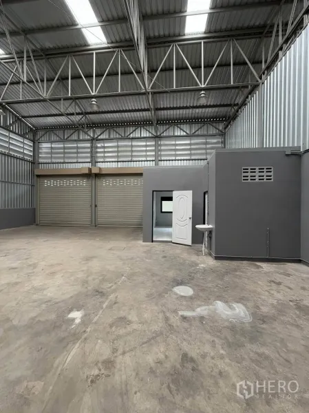 Warehouse for rent in Phra Pradaeng, Samut Prakan - Warehouse interior with high truss roof, concrete floor, two roller shutters, and an internal office room with a sink.