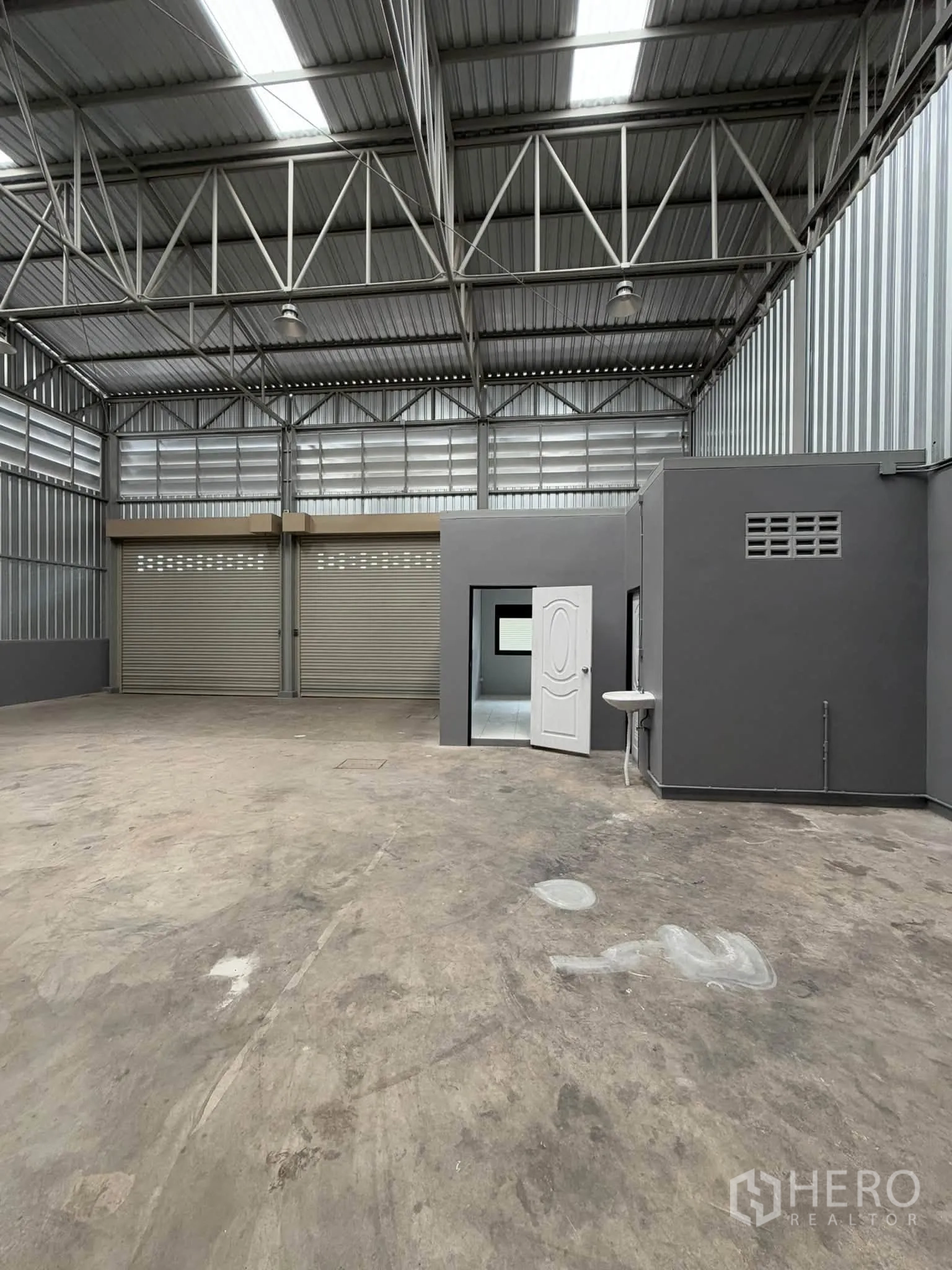 Warehouse for rent in Phra Pradaeng, Samut Prakan - Warehouse interior with high truss roof, concrete floor, two roller shutters, and an internal office room with a sink.