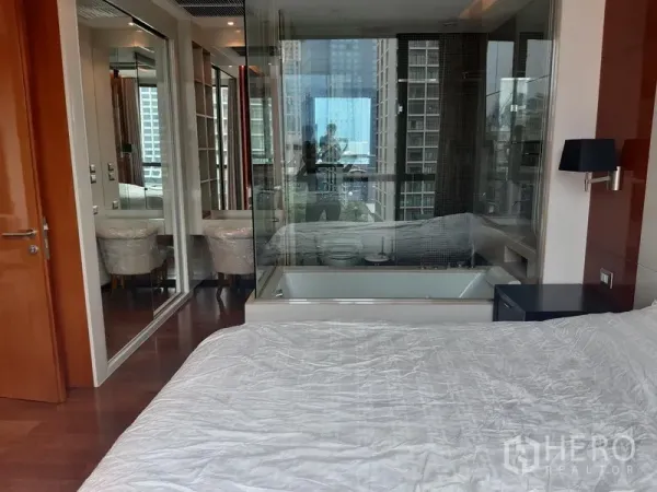 Condominium for rent in Khlong Toei, Bangkok - Bedroom with hardwood floors and a glass-partitioned bathroom featuring a soaking tub and city skyline outlook.
