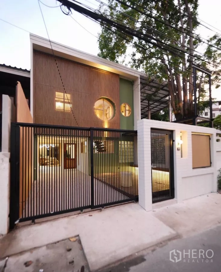 Townhouse for rent in Watthana, Bangkok - Japanese-style townhouse facade at dusk with circular windows, vertical wood slats and a black metal gate lit warmly.