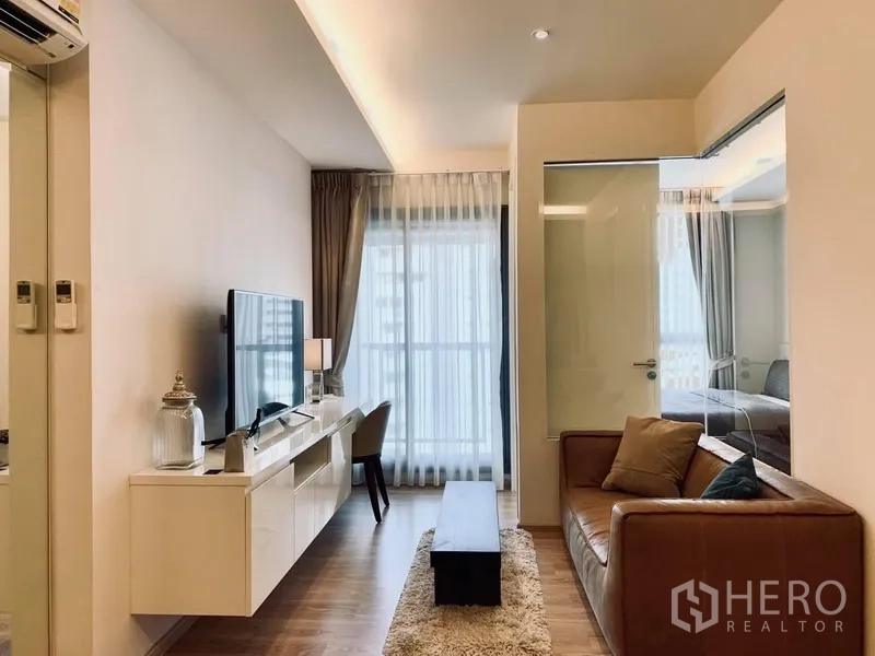 公寓 出租在Watthana, Bangkok - 明亮客厅，皮沙发、电视柜与落地窗，玻璃隔断通往卧室。