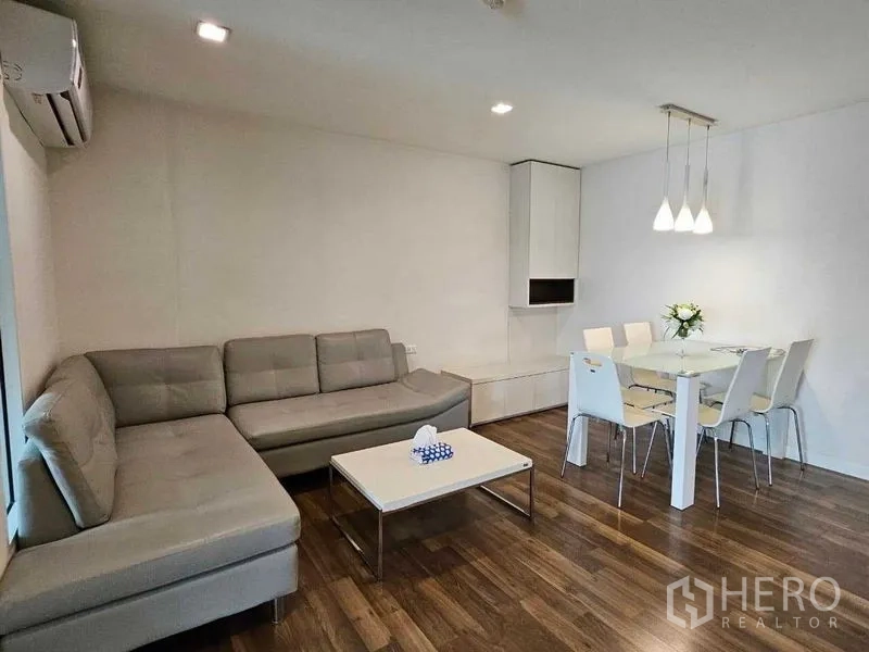 Condominium for sale in Watthana, Bangkok - Another view of the living room showing gray sectional sofa, dining table, and ceiling lights.