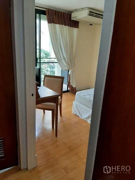 Apartment for rent in Watthana, Bangkok - Cozy bedroom with a study desk by the balcony, wood floors, and draped curtains.