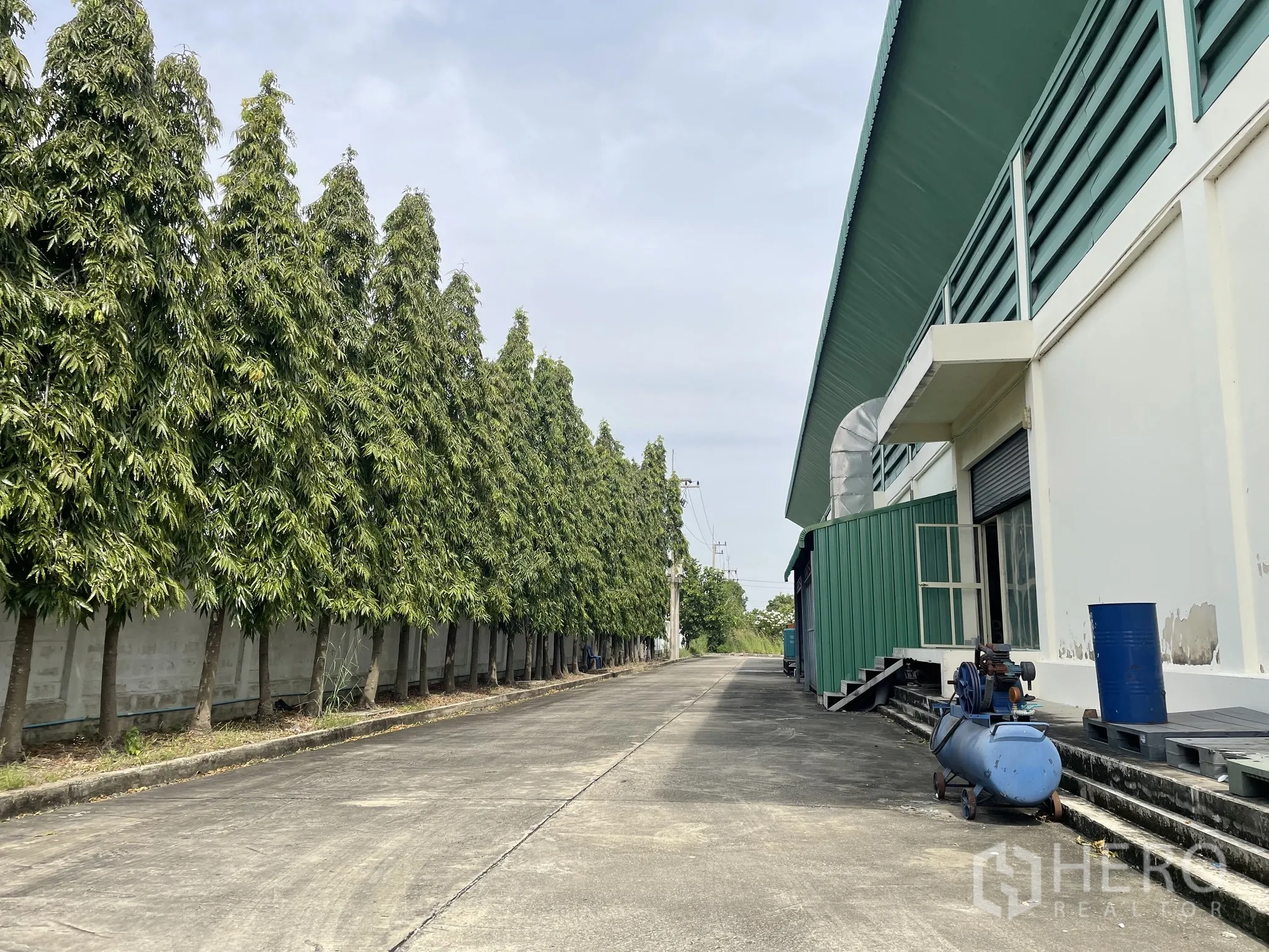 Warehouse for sale in Lat Lum Kaeo, Pathum Thani - Rear service road with ventilation ducts and equipment along the wall, shaded by tall trees.
