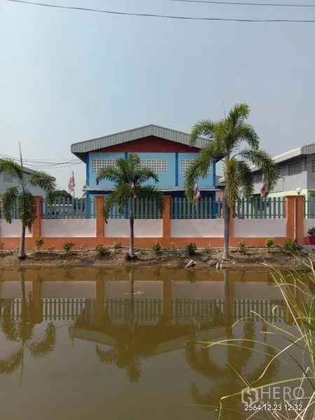 Factory for rent in Lat Lum Kaeo, Pathum Thani - Factory facade behind an orange-and-white perimeter wall with palm trees by a canal.