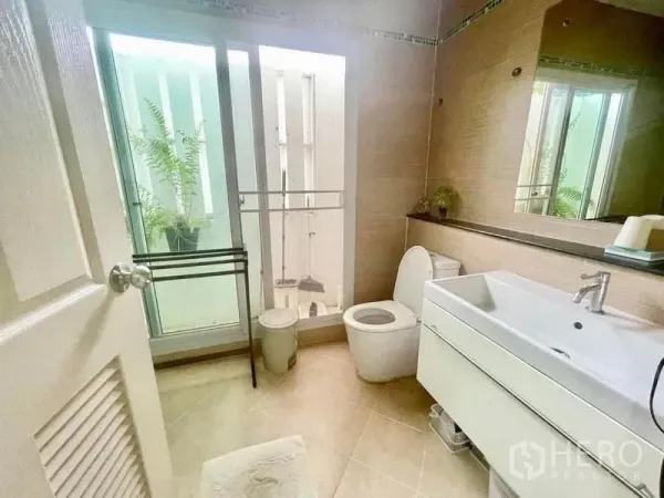 Detached House for rent - Bathroom with beige tiles, wide mirror, white vanity and toilet, opening to a lightwell with plants.