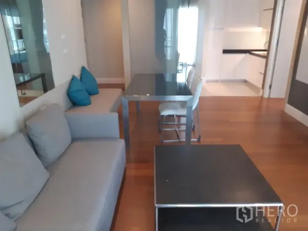 Condominium for rent or sale in Khlong Toei, Bangkok - Open-plan lounge with gray sofa and black dining table leading toward a sleek white kitchen on hardwood floors.