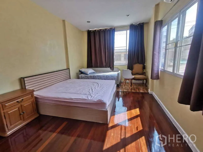 Townhouse for rent in Chatuchak, Bangkok - Sunlit bedroom with hardwood floors, double bed, side table and large windows with curtains.