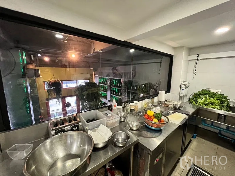 Shophouse for rent in Watthana, Bangkok - Stainless-steel prep room with a pass-through window overlooking a double-height dining space with hanging plants.