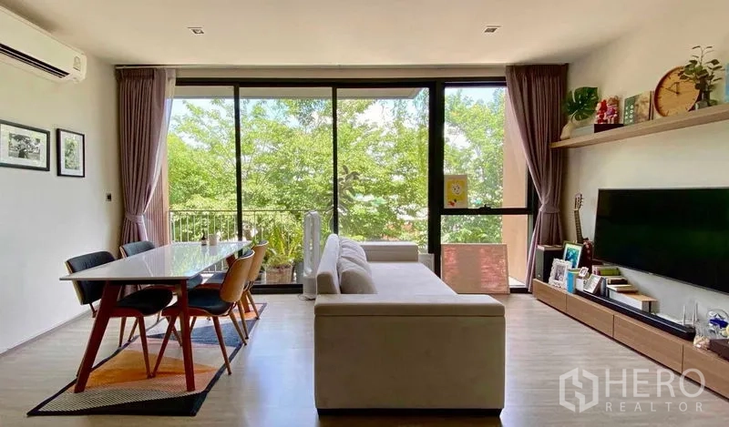 คอนโด ขายในWatthana, Bangkok - โถงนั่งเล่นและรับประทานอาหารแบบโอเพ่นแพลน พร้อมโซฟาและโต๊ะ มองออกสู่กระจกเต็มบานและระเบียงวิวต้นไม้