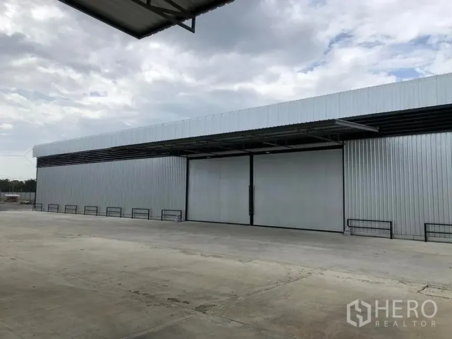 货栈 出租在Pathum Thani - 大型金属外墙仓库正面，超宽推拉门与宽阔混凝土装卸场，天空多云。