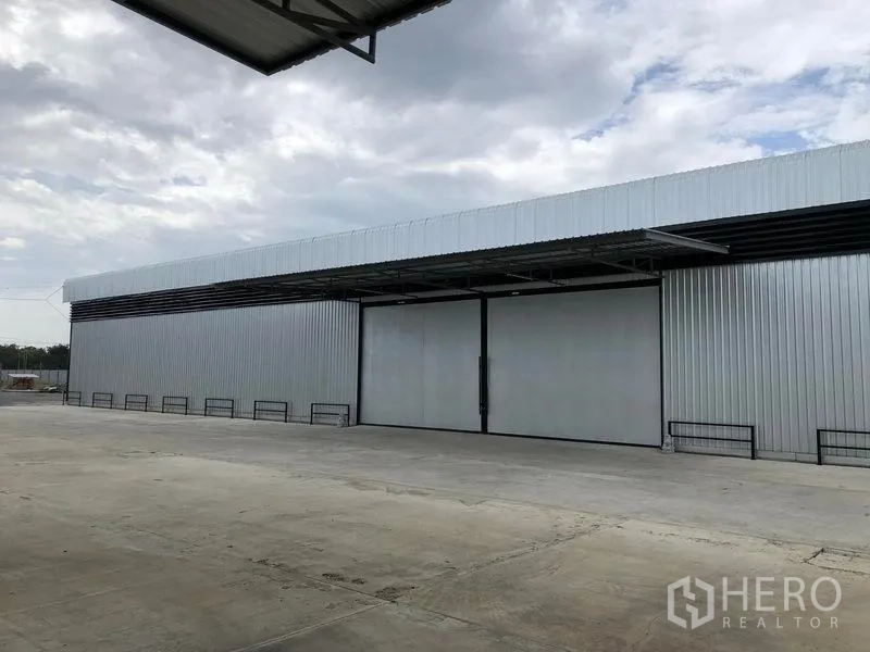 货栈 出租在Pathum Thani - 大型金属外墙仓库正面，超宽推拉门与宽阔混凝土装卸场，天空多云。