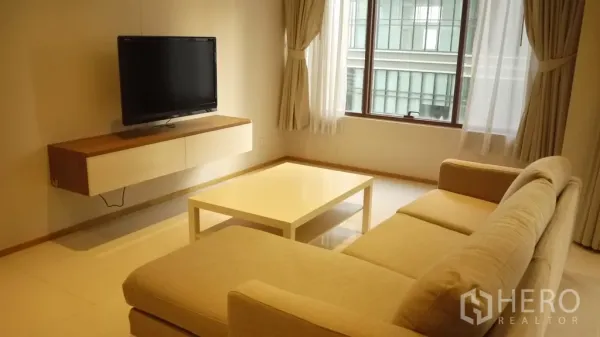Condominium for rent in Khlong Toei, Bangkok - Bright living room with beige sectional sofa, wall-mounted TV, coffee table and large curtained windows.