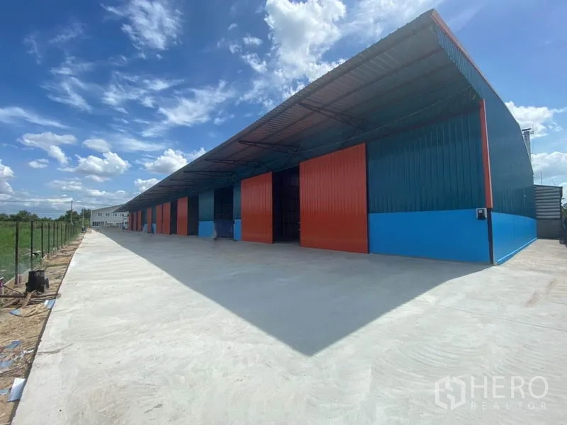 โรงงาน ให้เช่าในKhlong Luang, Pathum Thani - ภาพมุมกว้างด้านนอกโรงงาน ประตูสีแดงหลายช่อง และลานคอนกรีตกว้างท้องฟ้าแจ่มใส