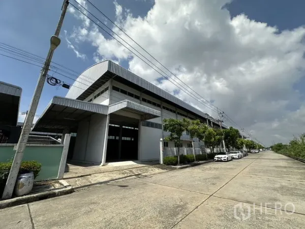 Factory for rent in Lam Luk Ka, Pathum Thani - Side view of large metal-clad factory building with tree-lined street and parking.