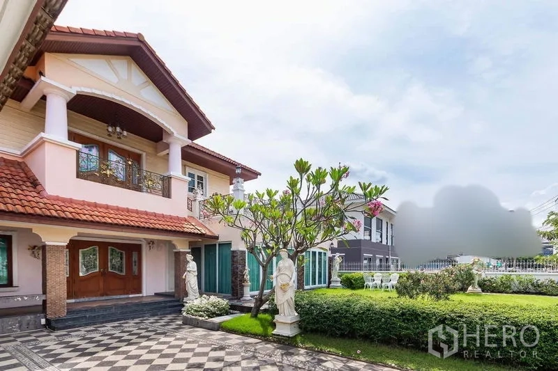 Detached House for rent or sale in Bang Khen, Bangkok - Grand entrance porch with double wooden doors, balcony columns, frangipani tree, and expansive front garden.