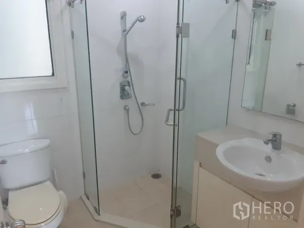 Condominium for rent in Khlong Toei, Bangkok - Clean white bathroom with glass-enclosed shower, vanity basin, and toilet by a window.