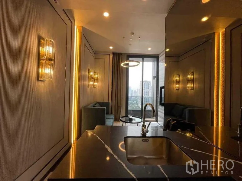 Condominium for rent in Khlong Toei, Bangkok - Open‑plan living room viewed from a black marble kitchen island, with ring pendant lighting and floor‑to‑ceiling window to a city‑view balcony.