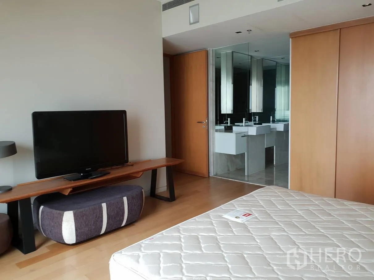 Condominium for rent in Sathon, Bangkok - Bedroom corner with TV console and an ensuite bathroom featuring double vanities behind a glass partition.
