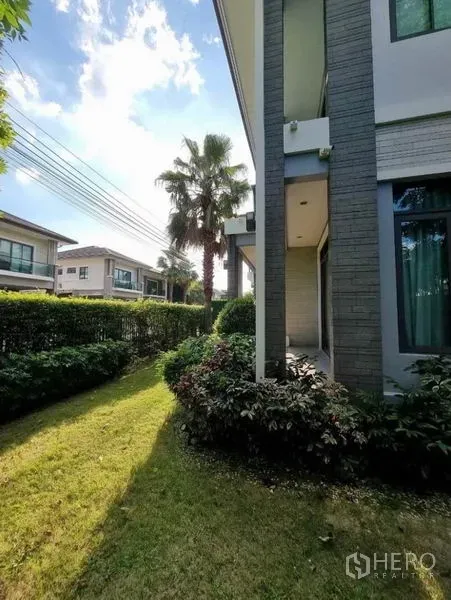 独立式住宅 出售在Suan Luang, Bangkok - 房屋侧边转角花园，低矮绿篱与棕榈树，可见邻里住宅。
