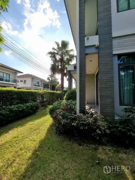 独立式住宅 出售在Suan Luang, Bangkok - 房屋侧边转角花园，低矮绿篱与棕榈树，可见邻里住宅。