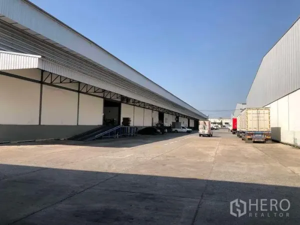 Warehouse for rent in Mueang Samut Prakan, Samut Prakan - Large warehouse complex with covered loading bays, wide driveway and truck parking under a clear sky.