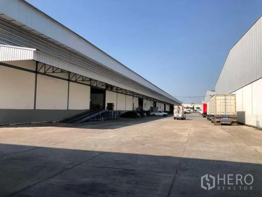 Warehouse for rent in Mueang Samut Prakan, Samut Prakan - Large warehouse complex with covered loading bays, wide driveway and truck parking under a clear sky.