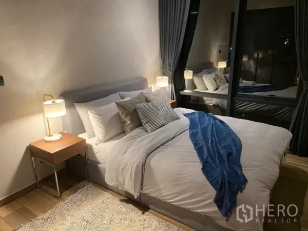 Condominium for rent or sale in Watthana, Bangkok - Primary bedroom with upholstered bed, bedside lamps, and curtain‑lined floor‑to‑ceiling windows.