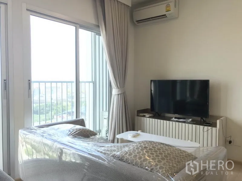 Condominium for rent in Huai Khwang, Bangkok - Living area with balcony doors, city view, TV cabinet and wall‑mounted air conditioner.