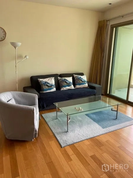 Condominium for rent in Khlong Toei, Bangkok - Cozy living room with sofa, armchair, glass coffee table, and sliding doors to the balcony.