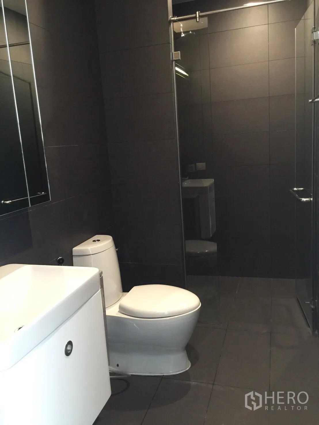 Condominium for sale in Pathum Wan, Bangkok - Second bathroom with large mirror, white vanity, and glass-enclosed shower against charcoal tiles.