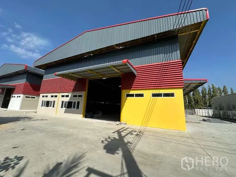 货栈 出租在Lam Luk Ka, Pathum Thani - 仓库外观，红黄配色的大型卷门与雨棚，宽阔混凝土场地与蓝天，位于Lam Luk Ka。