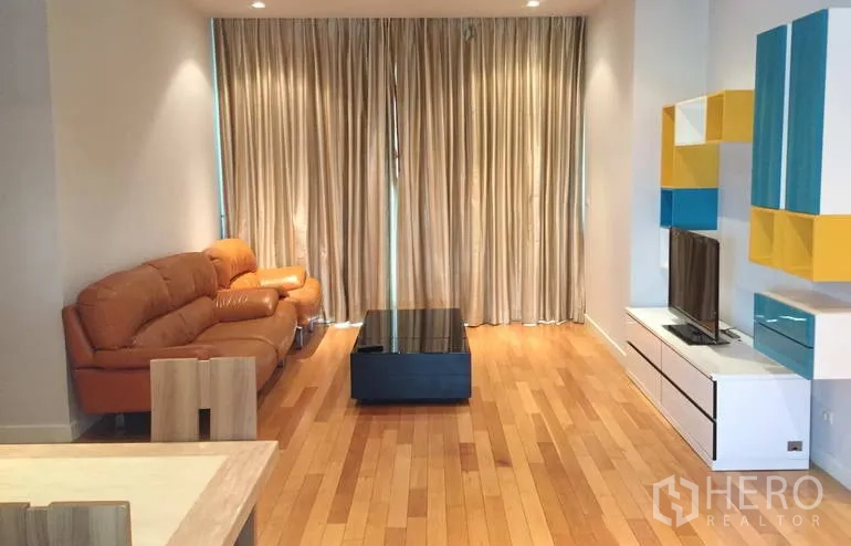Condominium for sale in Khlong Toei, Bangkok - Bright living room with tan leather sofa, floor-to-ceiling curtains, TV wall and hardwood floors.