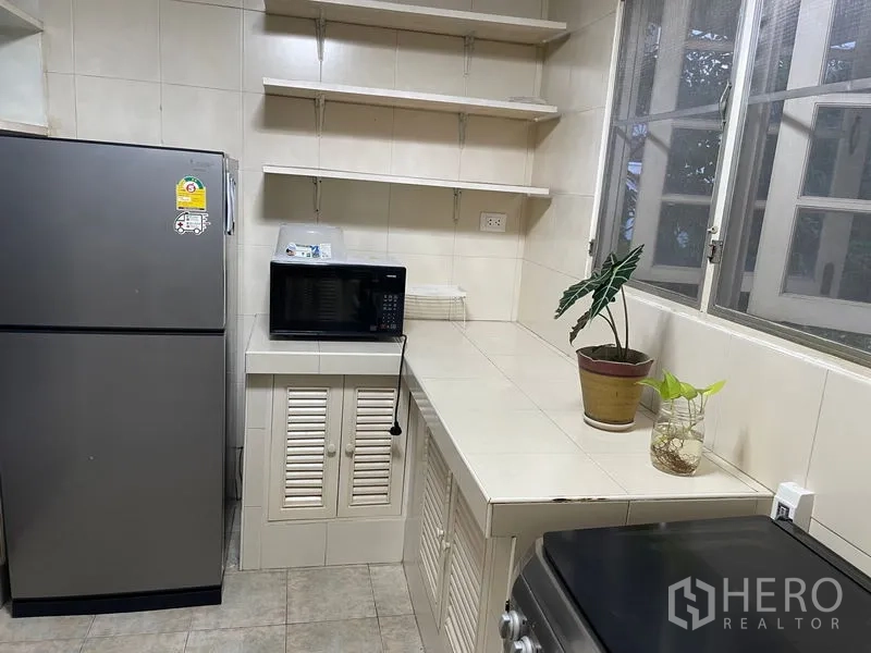 Apartment for rent in Watthana, Bangkok - Compact tiled kitchen with large fridge, microwave on the counter, open shelves and a window.