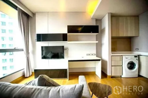Condominium for rent in Khlong Toei, Bangkok - Living area with built-in TV wall, work desk and kitchenette featuring an in-unit washing machine.