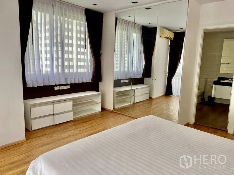 Condominium for rent in Khlong Toei, Bangkok - Bedroom with mirrored wardrobe, low cabinets beneath windows and en-suite bathroom door.