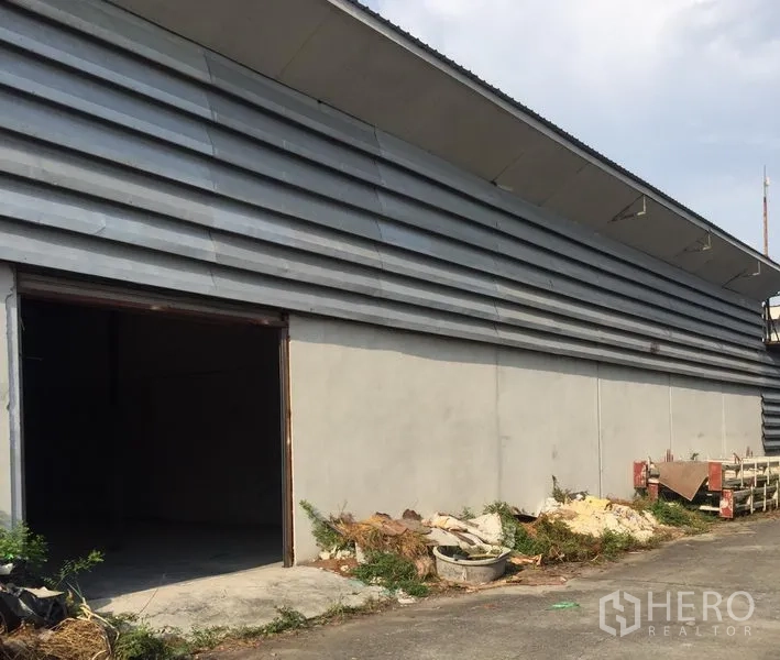 Warehouse for rent in Khlong Luang, Pathum Thani - Warehouse exterior with metal cladding, open roller door, and side yard for loading and equipment.